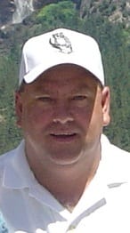 A man in white shirt and hat standing next to bushes.